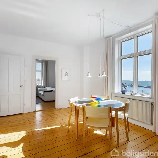 Rundt spisebord med fire stole står i en lys stue med trægulv. Store vinduer med udsigt over havet og dør til et tilstødende soveværelse ses i baggrunden.