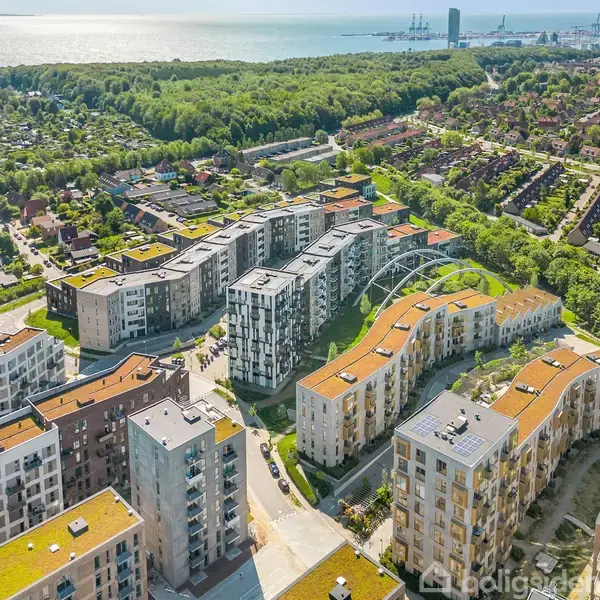 Bygninger med grønne tage står tæt arrangeret omkring en central vej. Omkringliggende områder omfatter træer, huse og kystlinje mod en fjord eller hav.