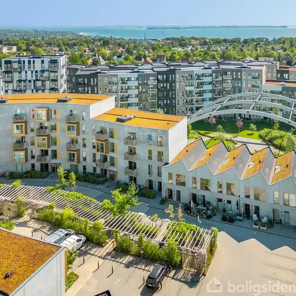Bygning med grønne tage står stille blandt flere moderne lejlighedskomplekser i et grønt boligområde. I baggrunden ses flere huse og en skov, havet kan også skimtes.