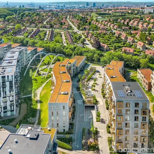Moderne lejlighedsbygninger med buede tage ligger i et grønt område med stisystemer. Omgivet af ældre huse i baggrunden, mens man kan ane byens skyline og industri i horisonten.
