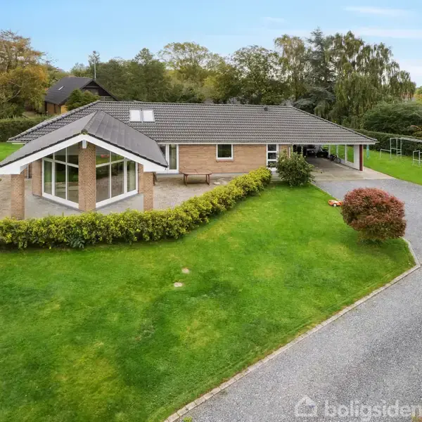 Et enfamiliehus med grå tagsten omgivet af velplejet græsplæne og hække. En grusvej fører til carporten på højre side.