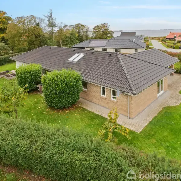 En enkeltstående, murstensvilla med gråt tag står på en velplejet græsplæne omgivet af hække. I baggrunden ses flere huse og vandområde, der skaber en naturskøn atmosfære.