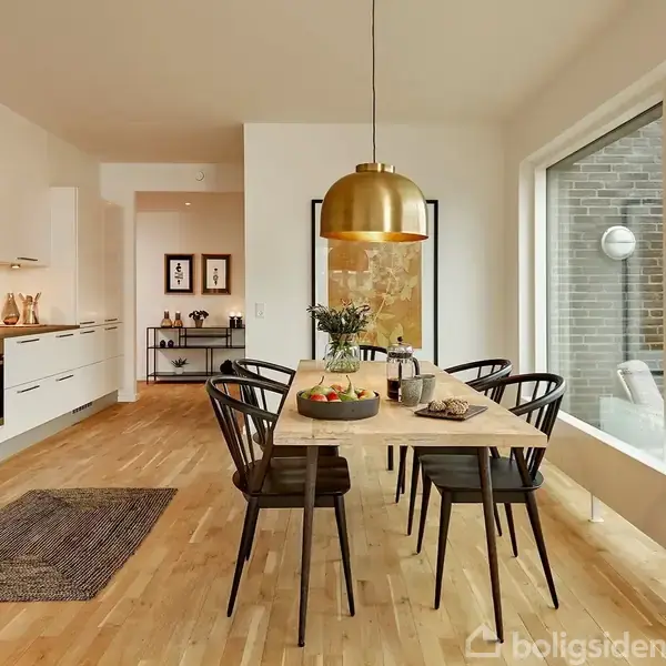 Spisebord af træ med sorte stole står under en gylden loftslampe. I køkkenet i baggrunden er hvide skabe og en ovn. Store vinduer giver udsigt til en lejlighedsbygning.