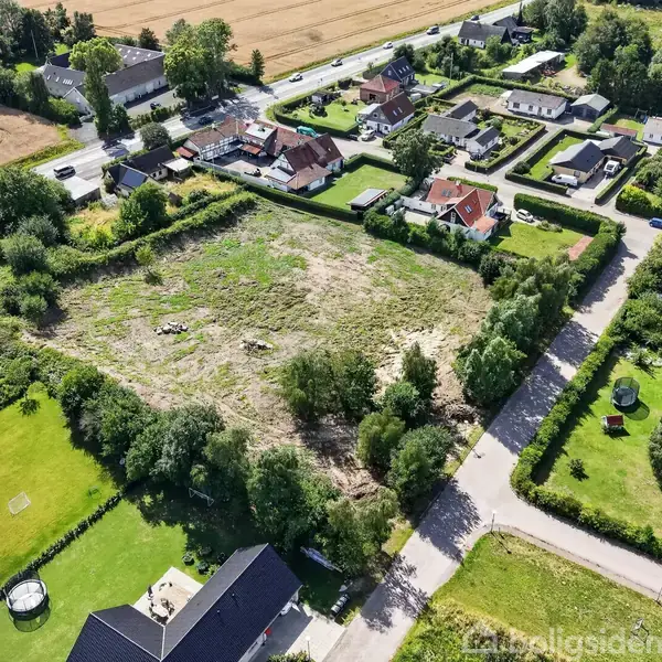Et grønt område omgivet af huse og marker; midten viser en åben, ufærdig grund. Træer og buske omkranser stedet, der grænser op til villaveje og haver.