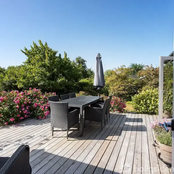 Terrasse med sorte kurvemøbler og parasol står i en have omkranset af blomsterbuske. Lyset er klart, og himlen er blå.