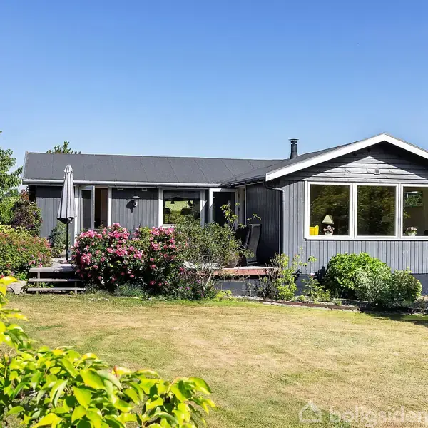 En gråmalet bungalow ligger i en frodig have med blomstrende buske og frugtbare træer. Huset har store vinduer og en terrasse, der skaber en hyggelig atmosfære i naturen.