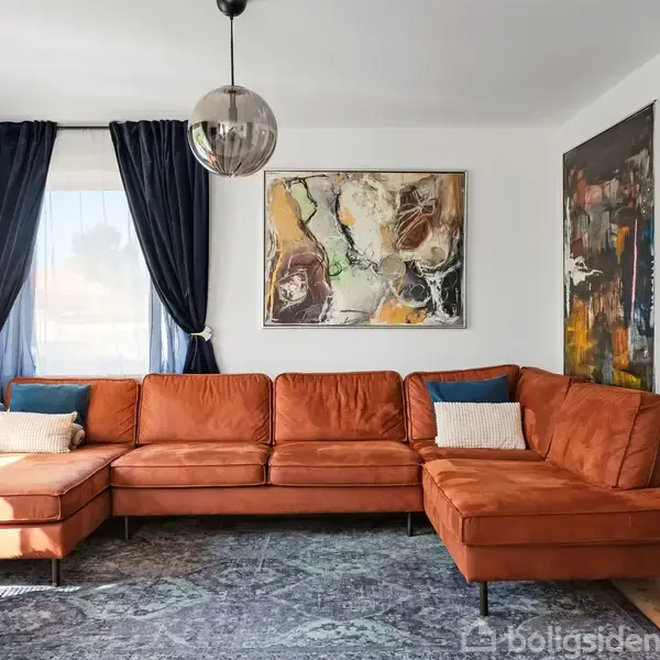 En orange sofa står centralt i en stue med et stort kunstmaleri på væggen og blå gardiner ved vinduet. En rund lampe hænger ned fra loftet.