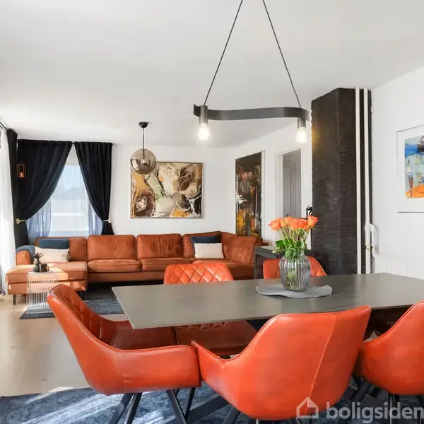 En moderne, lys stue har en stor orange sofa og matchende spisebordsstole. Et langbord med en vase med blomster på står centralt. Væggene er dekoreret med kunstværker.