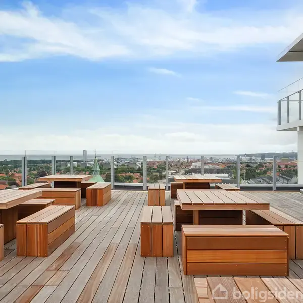 Tagterrasse med træmøbler arrangeret til siddepladser, ingen personer til stede. I baggrunden ses udsigt over byen med himmel og skyer. Ingen tekst findes på selve billedet.