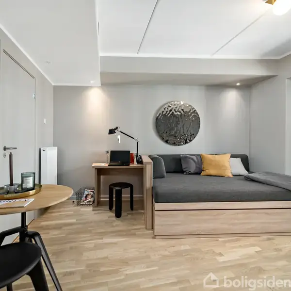 Værelse med sovesofa, skrivebord og stol, set i et moderne lejlighedsområde. Rummet har lyse trægulve og adgang til en balkon med udsigt over byen.