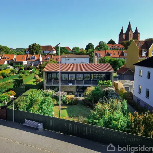 En moderne villa med store vinduer står i en velplejet have. Naboer er tæt placeret, og en kirke med to spir ses i baggrunden.