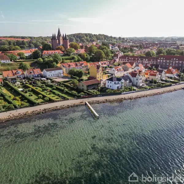 Kystby med farverige huse langs vandet og velplejede haver. En kirke med spir dominerer baggrunden, omgivet af skov og by. Billedet bemærker detaljerede haveanlæg og en badebro ved vandet.