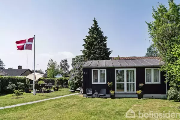 Et sort sommerhus med hvidt dørparti står i en grøn have. Et dansk flag vajer på en flagstang til venstre. Huset har nummeret "44" ved døren.