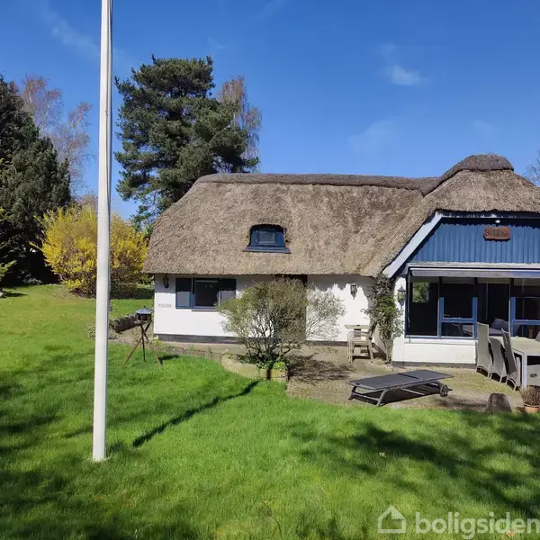 Et bindingsværkshus med stråtag står stille i en grøn have med træer og buske i baggrunden. En flagstang og havemøbler er synlige omkring huset.
