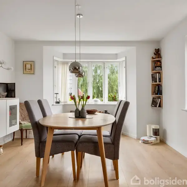 Et rundt spisebord med fire stole står i midten af en lys stue. Der er en vase med blomster på bordet. Et stort vindue lader naturligt lys strømme ind.