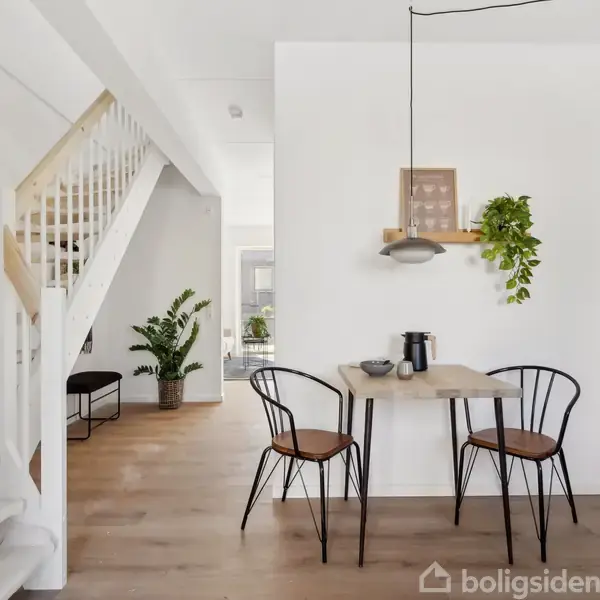 Et lille spisebord med to stole står i et moderne køkken. En plante og en lampe hænger over bordet. Trappe og grønne planter ses i baggrunden.