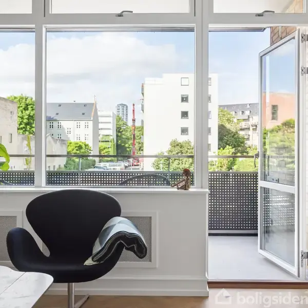 En moderne stue har en sort stol ved et åbent vindue, der fører ud til en altan. Grønne planter og klare glasruder giver udsigt til bygninger og træer udenfor.
