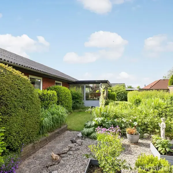 En velplejet have med grønne buske og blomster omgiver et hus med røde mursten. En stensti fører gennem haven, der indeholder en lille statue og blomsterbede.