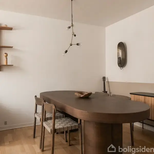 Et ovalt træbord står i en stue omgivet af stole. Til venstre hænger en hylde med keramik, og til højre ses en kommode med vaser. En lampe hænger fra loftet.