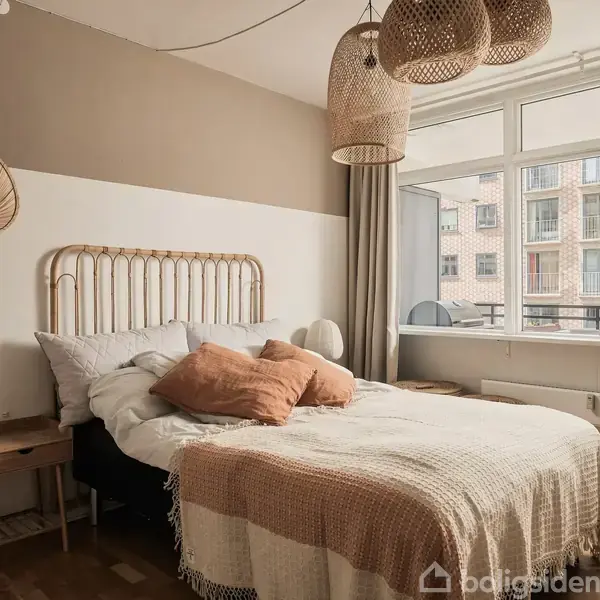 En seng med hvide og beige puder står i et lyst soveværelse med træmøbler. Vægpynterne inkluderer rattan-elementer, og store vinduer viser en udsigt til nabobygninger.