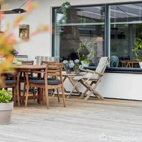Et træbord og stole står på en træterrasse foran et hus. Omkring bordet ses planter i krukker, og store vinduer giver udsigt til indretningen indenfor.