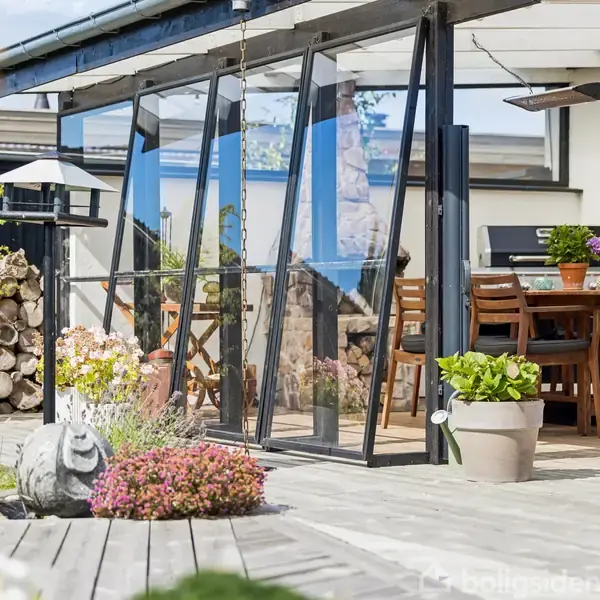 En overdækket terrasse med glasvægge. Træmøbler står under tag, omringet af blomster og planter. Stabler af brænde og en udendørs lampe ses i siden, skaber en hyggelig atmosfære.