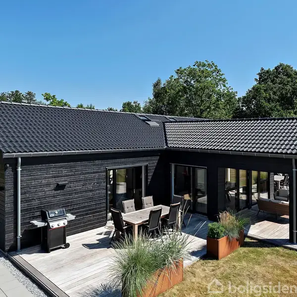 Sort træhus med terrasse, placeret i en grøn og frodig have med store træer i baggrunden. Terrassen har udendørs møbler og en grill, ideel til udendørs ophold.