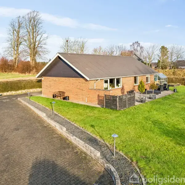 En lang, en-etagers murstenshus med sort tag står langs en hæk. Der er flisebelagte stier omkring huset, placeret i et åbent, grønt område med træer i baggrunden.