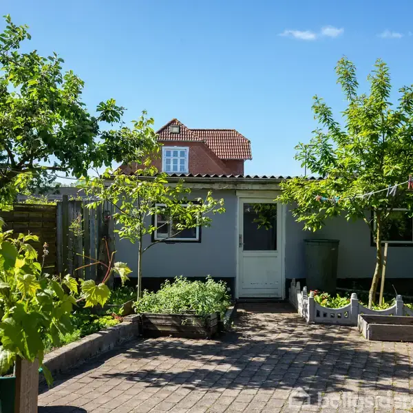 Et lille hus med en hvid dør ligger i en have med forskellige træer og plantebede. Brostenene leder op til indgangen, omgivet af grønne planter i en solrig omgivelse.