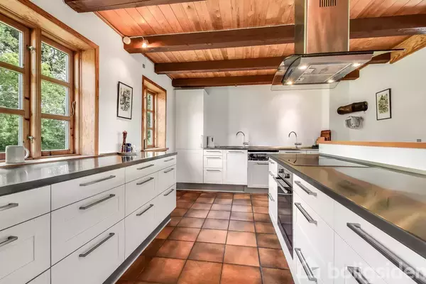 Køkken med hvide skabe og metaloverflader. Ovne indbygget i skabene. Terrakotta-fliser på gulvet. Vinduer viser grøn bevoksning udenfor. Træloft og synlige bjælker skaber en rustik atmosfære.
