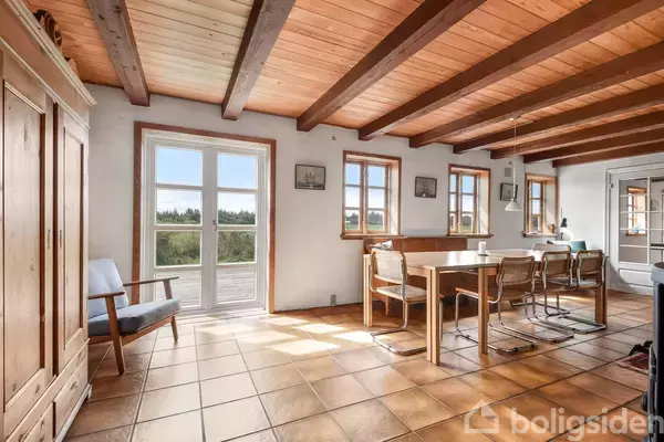 Træbord med stole er placeret i en spisestue med træloftsbjælker og klinkegulv. Store vinduer mod naturen lader lys strømme ind.