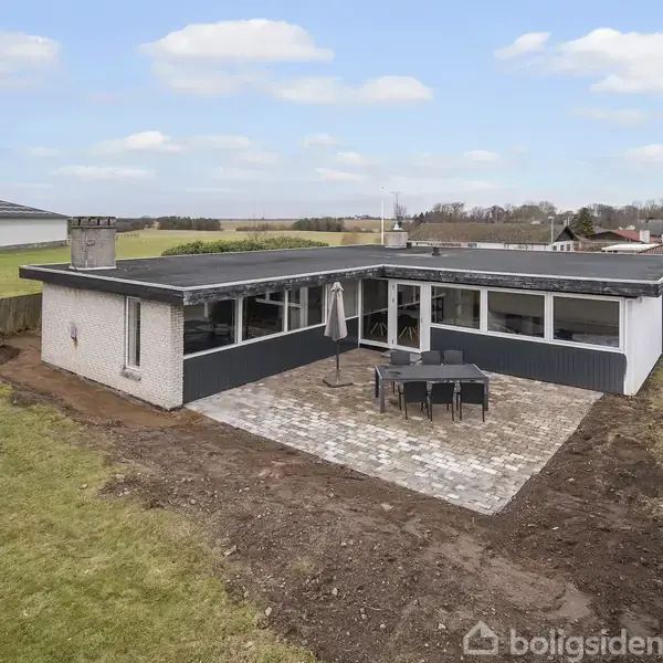 En moderne bungalow står på en stor grund; en terrasse pryder forsiden med bord og stole. Omkring er der en græsplæne og tætliggende hække.