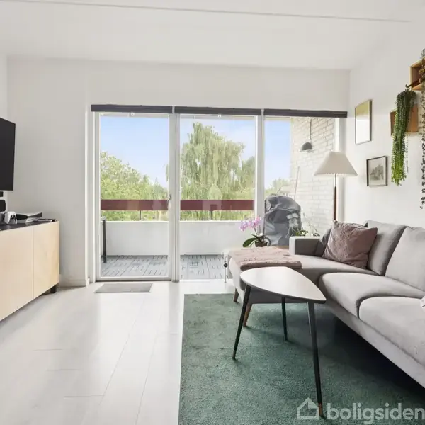 Sofa står i en stue ved siden af en glasdør, der fører ud til en balkon. Rummet er lyst med jay beige vægge, et sofabord og en reol med planter på væggen.