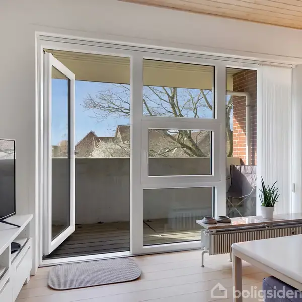 En stue med åben terrassedør leder ud til en balkon. Indenfor står et TV på et lavt bord, en sofa til højre, og en plante ved døren.