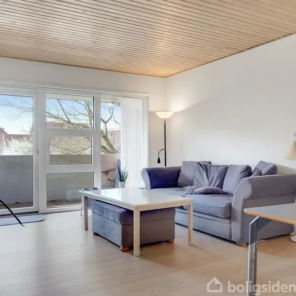 En grå sofa står i en stue ved siden af et hvidt sofabord. Rummet har trægulv, træloft og store vinduer, som viser en balkon med udsigt til yderlige træer og huse.