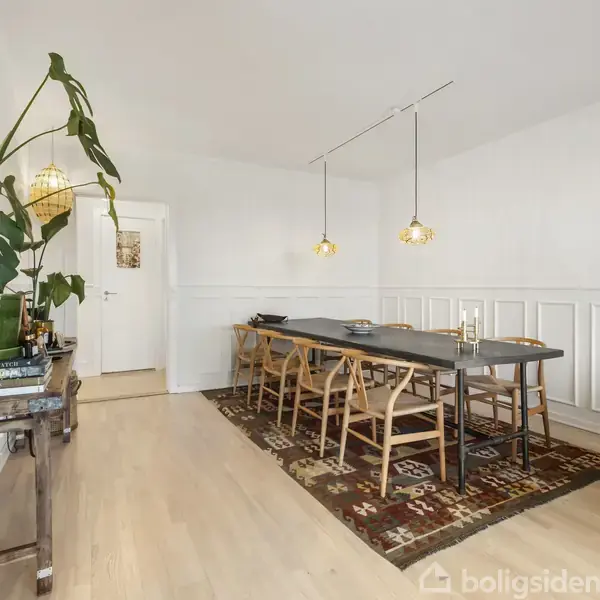 Langt spisebord med seks stole placeret i en lys spisestue. To lamper hænger over bordet. En stor plante står på et sidebord til venstre.