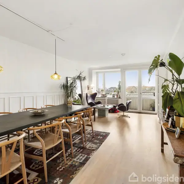 En spisestue med et langt mørkt spisebord omringet af stole. Rummet har to hængelamper, en farverig tæppe og store vinduer med udsigt. En plante står ved siden af et lille sofabord.