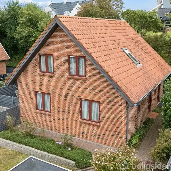Rødt murstenshus med skråt tegltag, omgivet af grønne buske og en græsplæne. Huset har flere vinduer og et loftvindue. Nabohuse og træer ses i baggrunden.