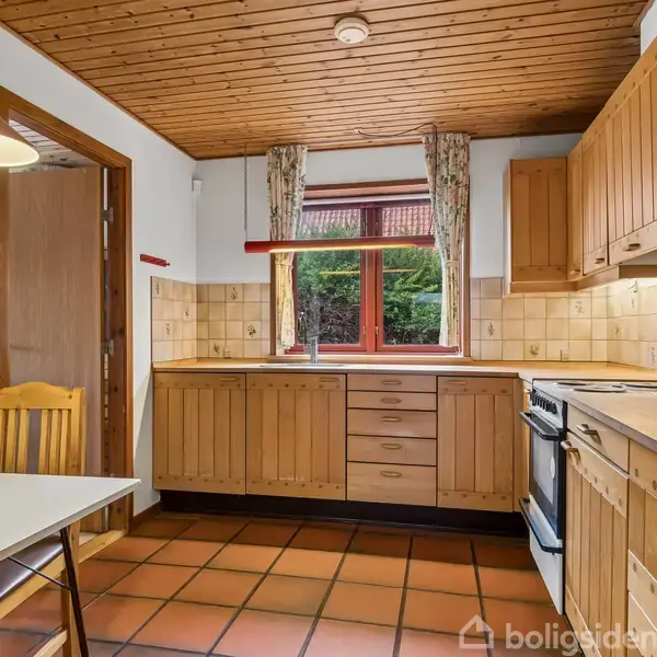 Køkken med træskabe og -loft. Bord med to stole står nær et vindue med udsigt til buske. Fliser på gulv og vægge skaber en rustik atmosfære.
