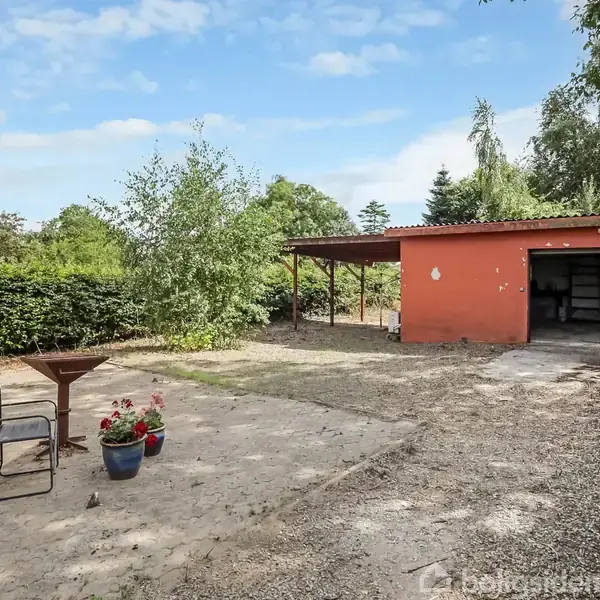 En rustik gårdsplads med et rødt skur til højre. Et bord og stol står ved siden af en blå blomsterkrukke. Omkring er der grønne buske og træer under en blå himmel.