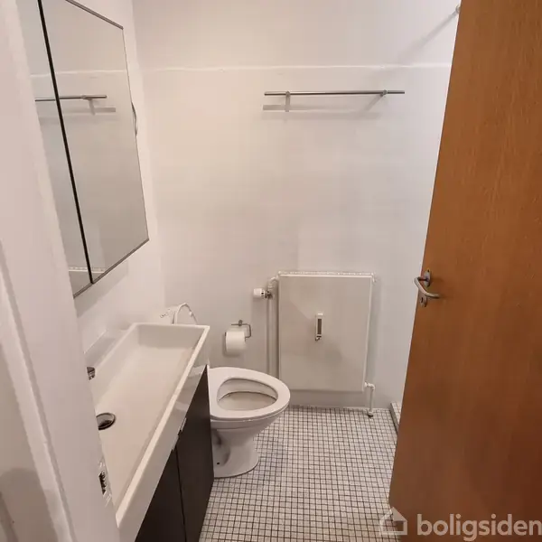 Toilet placeret midt i et lille badeværelse. På venstre side er en håndvask med spejl over. Højre væg har en åben dør og et lille opvarmningsapparat. Tekst ignoreret.