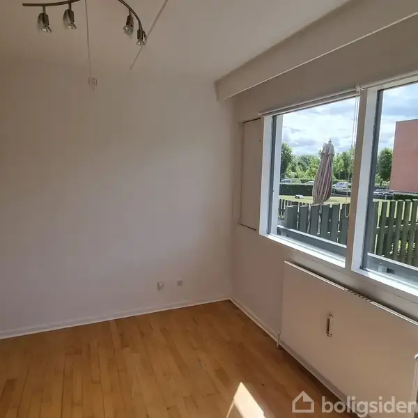 Et tomt værelse med trægulv, hvor sollys skinner fra et stort vindue til højre. Udenfor ses en træterrasse og bygninger. En loftlampe hænger over rummet.