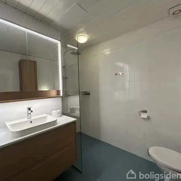Et moderne badeværelse med en vask på et træskab, indrammet spejl med lys, en bruser bag en glasvæg og et toilet i et lyst flisebelagt rum.