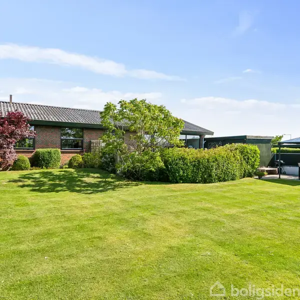 Hus står stille omringet af frodig have med klippede buske og grøn græsplæne under en klar blå himmel. Terrasse med overhæng på højre side.