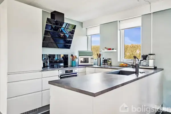 Et moderne køkken med hvide skabe og en grå køkkenø. Køkkenet har en indbygget ovn, kogeplader, og en moderne emhætte. Vinduer med udsigt til grønne buske giver naturligt lys.