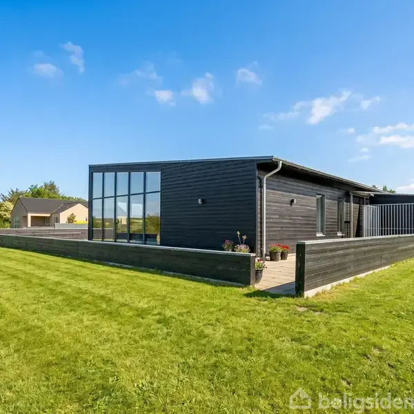Sort, moderne hus med store vinduer står på en velplejet græsplæne. Træterrasse omgiver huset. Solrig dag med klar blå himmel og få skyer.