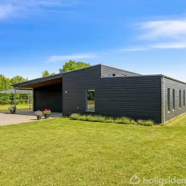 Et moderne, sort træhus står stille på en velplejet græsplæne omgivet af natur. Foran huset er en flisebelagt terrasse med planter i krukker.