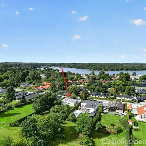 Hus med rød pil ovenover, omringet af træer og nabolag i grøn landsby nær en sø i naturligt landskab.