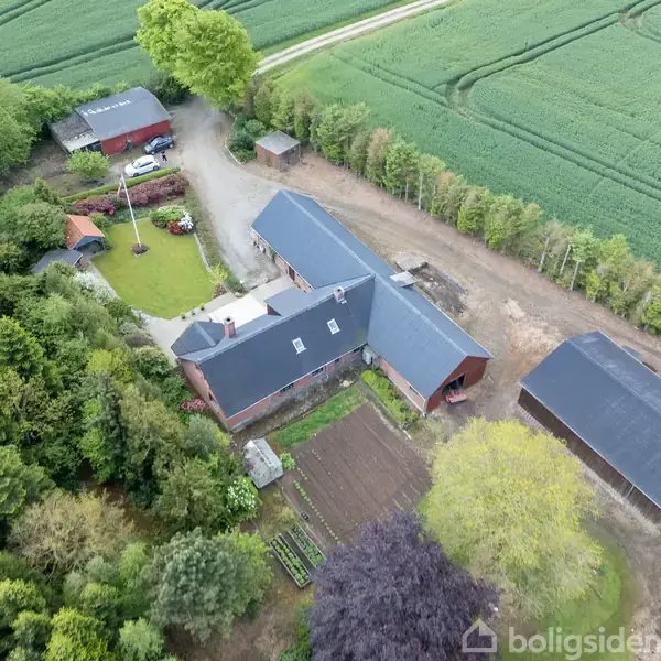 Gård beboelse med tre langstrakte bygninger omgivet af skov og marker; en have med flagstang foran huset og marker omkranset af træer i baggrunden.