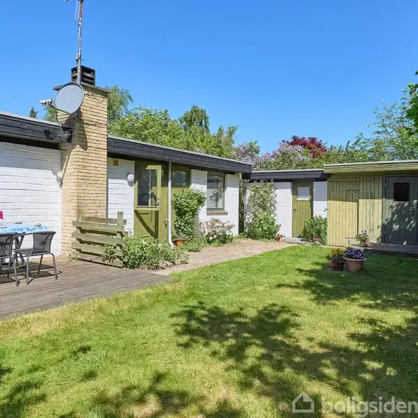 En hvid murstensbygning med en skorsten og grønne døre placeret i en have. En terrassestol står ved et bord med blåt dug. Græsplæne omgiver huset i solskinsvejr.